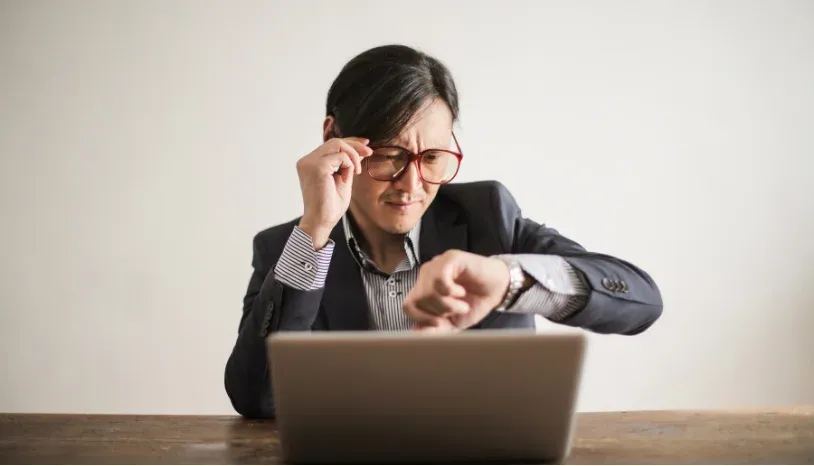 A man in a suit, sitting in front of his laptop, with his wrist raised while looking at this watch